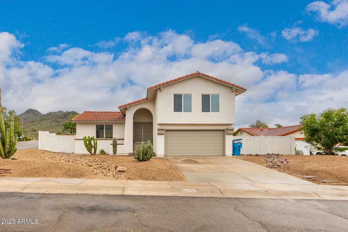 The exterior of a two-story house is presented, showcasing a light-colored facade and a red-tiled roof. A neat driveway leads to a garage, with desert landscaping featuring cacti and gravel. A mountain view is visible in the background under partly cloudy skies.