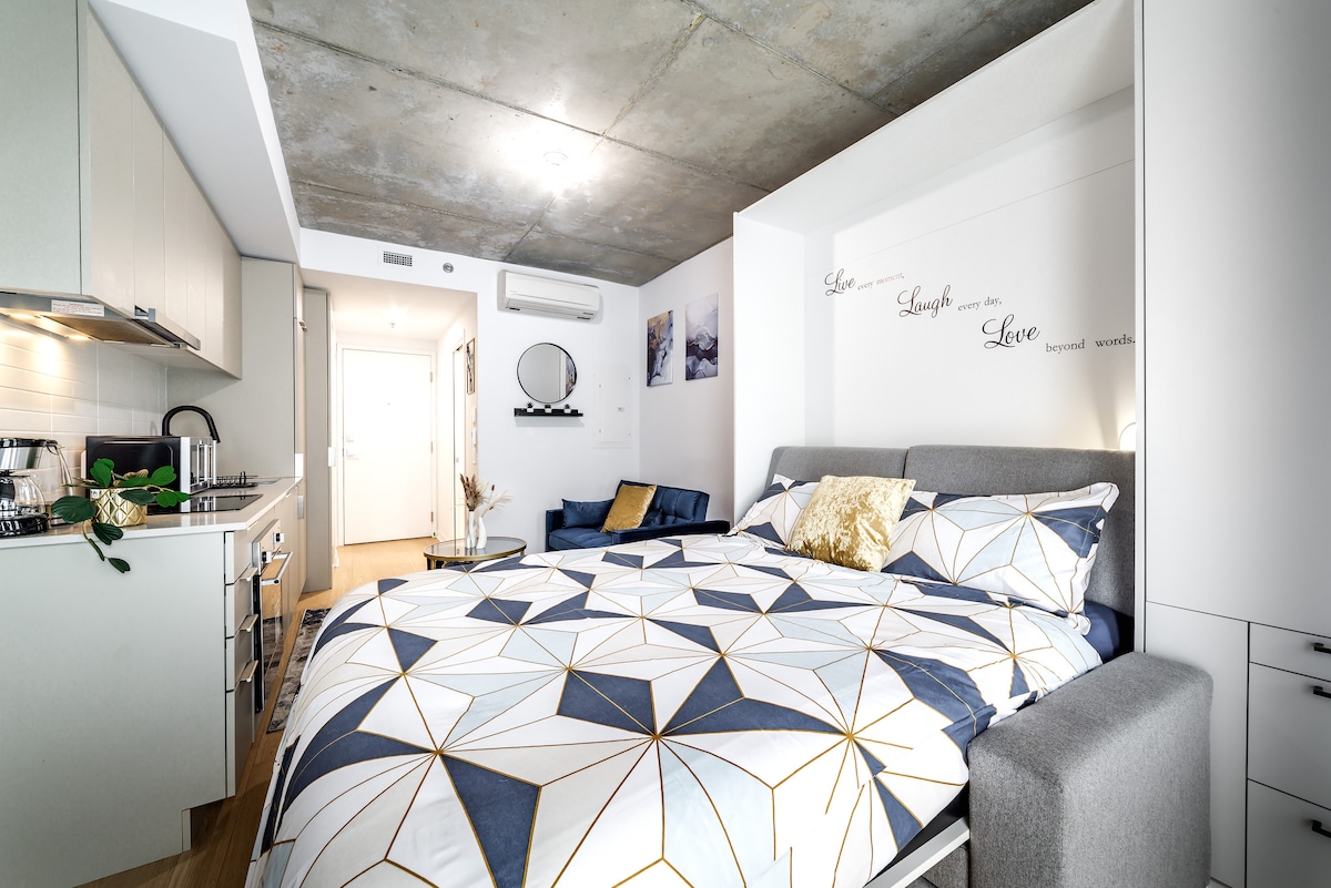 A modern studio space is showcased, featuring a spacious bed dressed in a geometric-patterned duvet. To one side, a compact kitchenette is visible, along with a small seating area furnished with a blue couch. Minimal wall art decorates the space, enhancing the contemporary design.