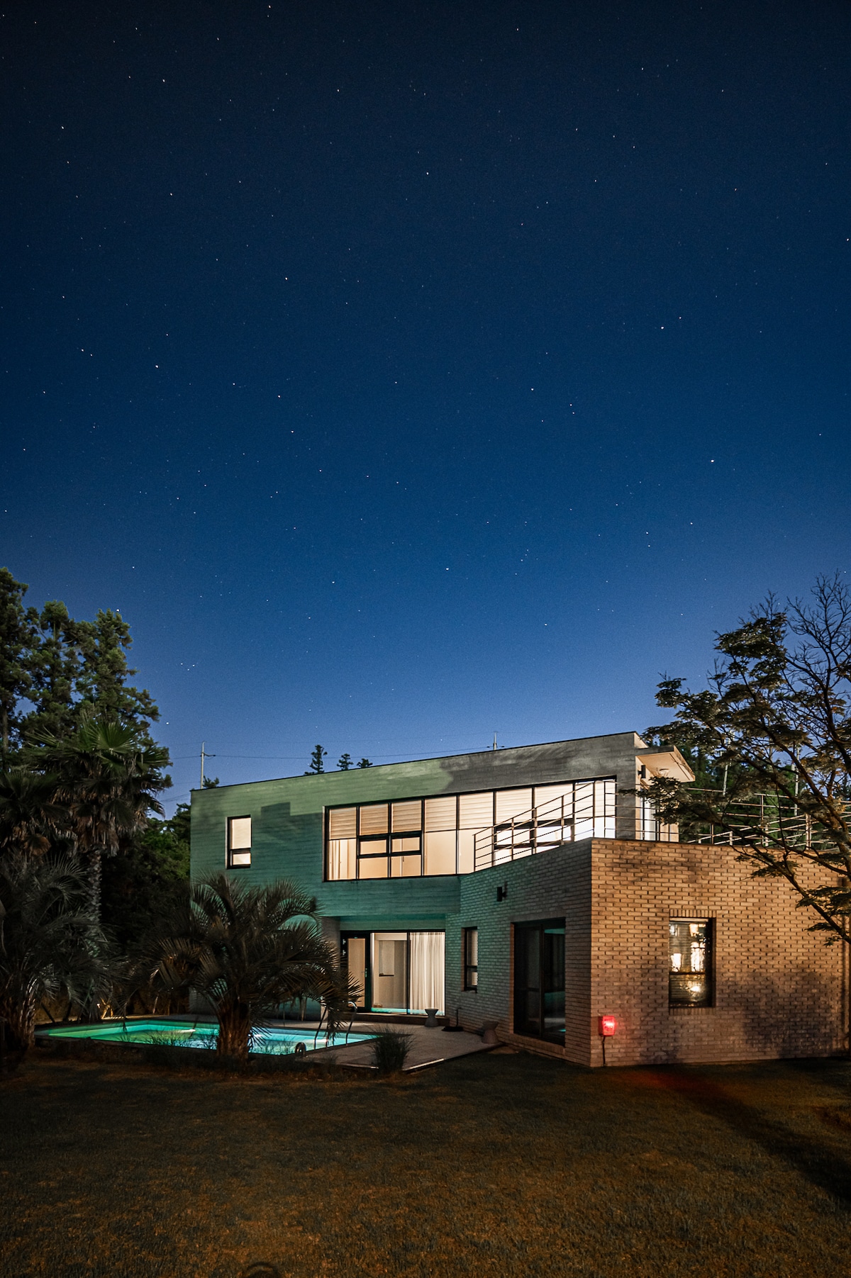 The modern two-story villa is illuminated under a starry sky, showcasing large glass windows that reflect the soft evening light. A private pool is visible along the side, surrounded by lush foliage, creating a serene outdoor space.