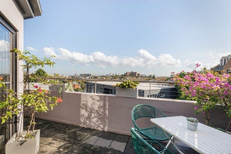A private balcony features a table and chairs surrounded by flowering plants. The view showcases the city skyline against a clear blue sky with scattered clouds, providing an inviting outdoor space for relaxation.