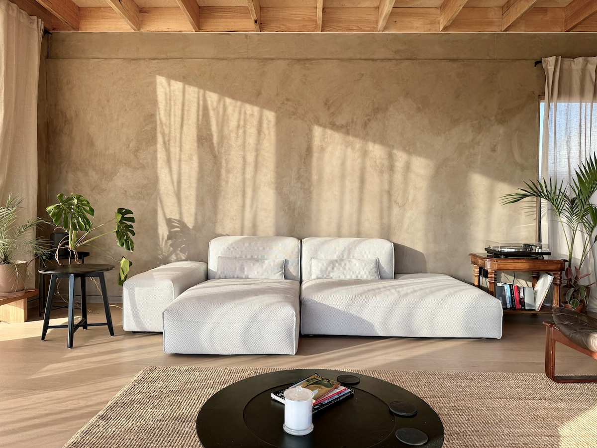 A minimalist living area features a spacious, light-colored sectional sofa set against a textured wall. Natural light casts gentle shadows across the room. A small black table sits in front, accompanied by potted plants, creating a serene and inviting space.