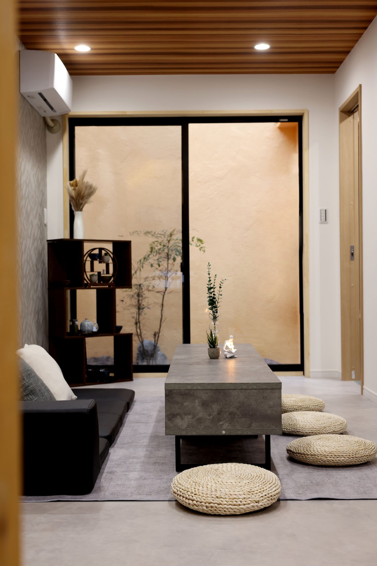 The living area is furnished with a dark sofa and a minimalistic concrete coffee table. Natural light enters through large glass doors, revealing a serene outdoor space with greenery. Woven floor cushions are arranged neatly on a soft area rug.