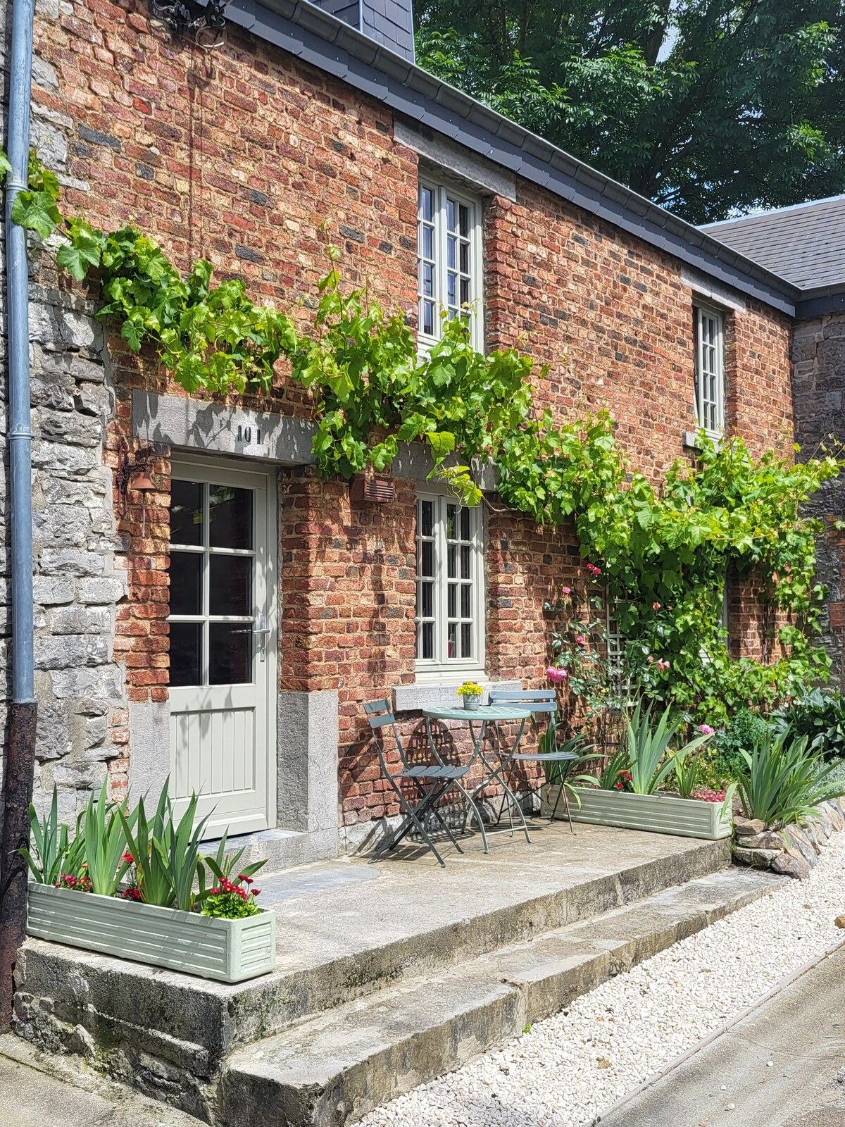 The exterior of the house is adorned with vibrant green vines against the rustic brick facade. A small table and two chairs are positioned on the stone steps, surrounded by neatly organized flower boxes. Soft sunlight highlights the inviting entrance.