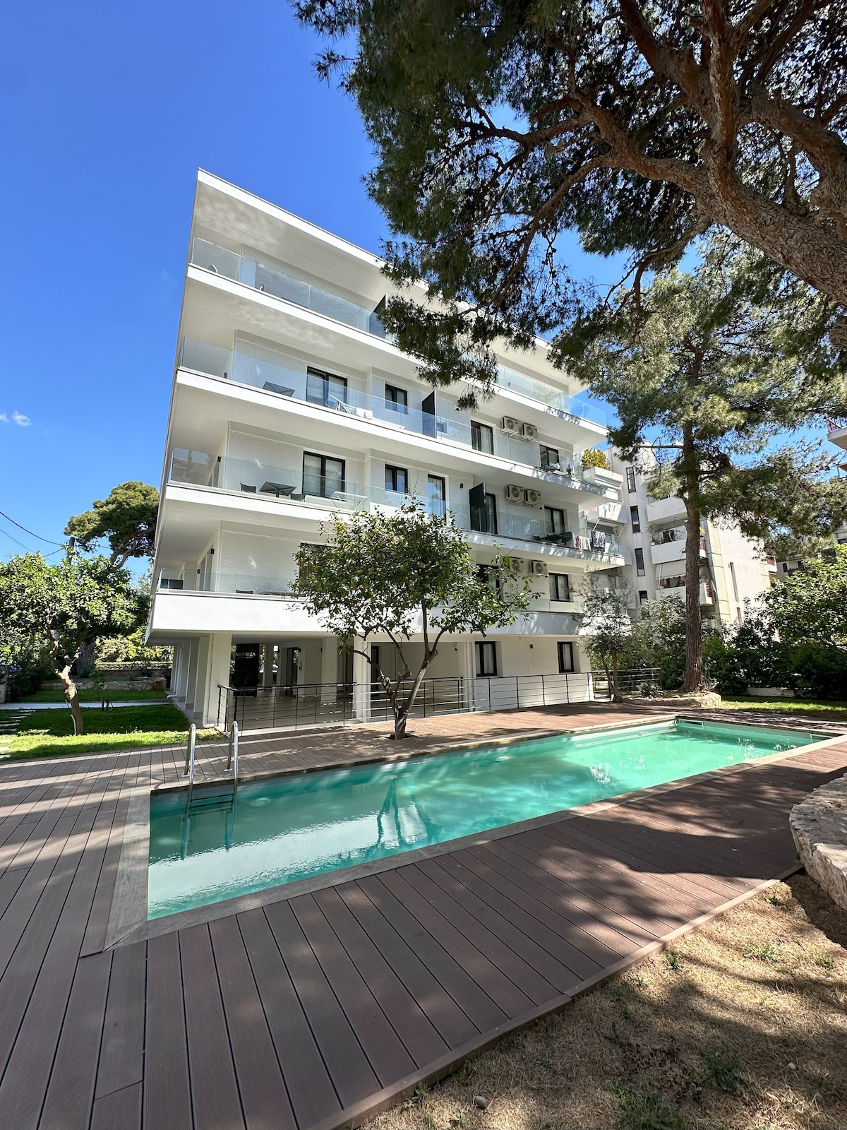 A modern multi-story building is surrounded by greenery, featuring a swimming pool with a sleek wooden deck. Various trees provide shade, enhancing the outdoor space. Large balconies are visible on each floor, promoting views of the garden area.
