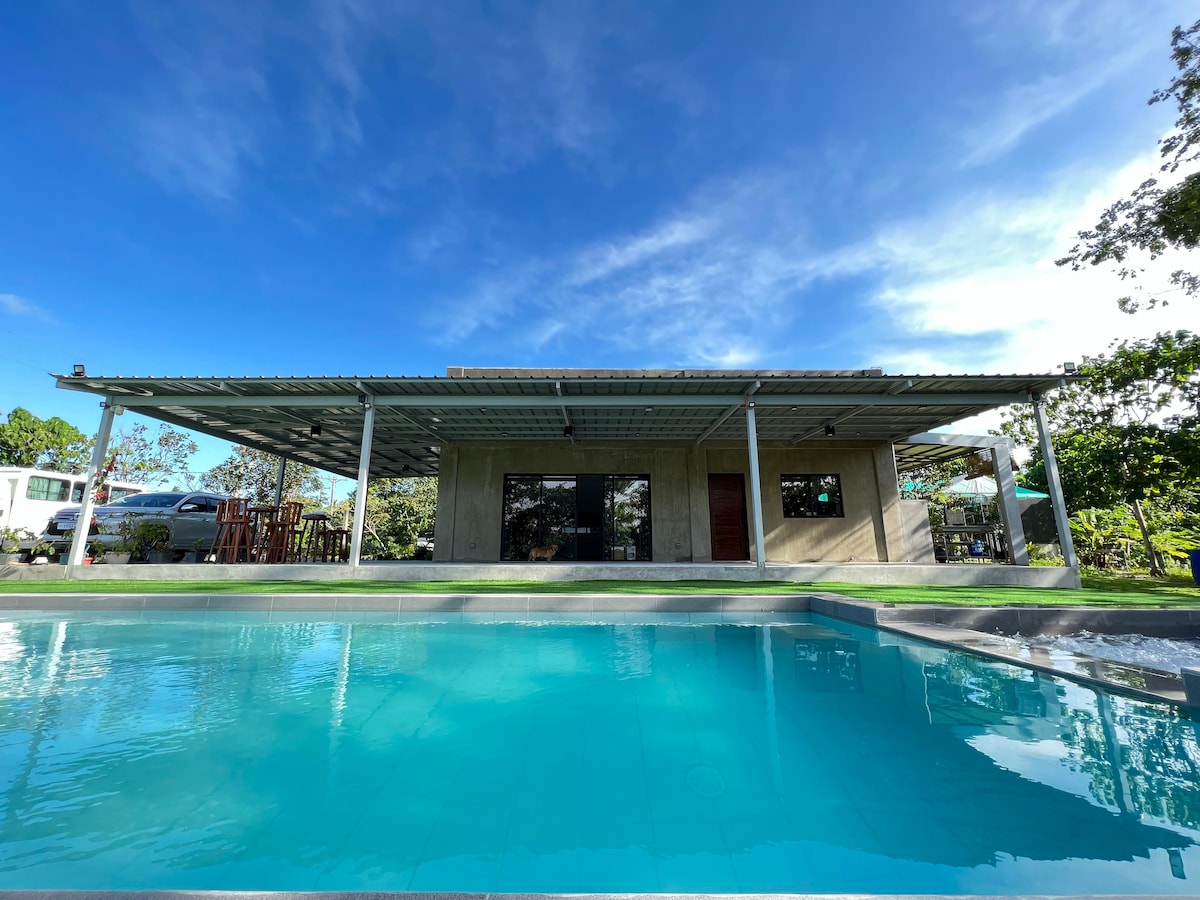 A modern home stands adjacent to a clear blue pool, with a spacious covered patio featuring large glass windows. The structure is surrounded by lush greenery, allowing an open view of the sky above. The setting offers a serene and inviting atmosphere.