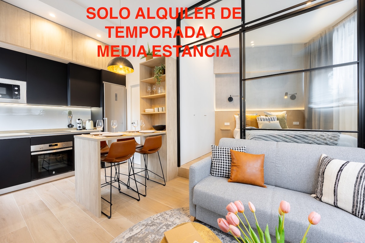 A modern living space features a cozy sofa, complemented by a decoratively folded orange throw and a vase with fresh pink tulips on the table. The open kitchen showcases sleek black cabinetry and modern appliances, while a warm light ambiance is created with stylish pendant lighting.