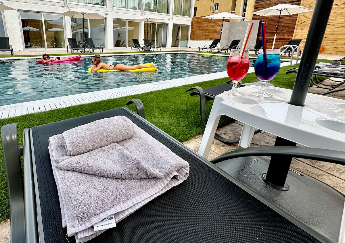 A pair of colorful cocktails sits on a table beside a lounger, with neatly folded towels placed atop it. The view includes guests relaxing on floats in a refreshing pool, surrounded by sun loungers and umbrellas, creating a serene poolside atmosphere.