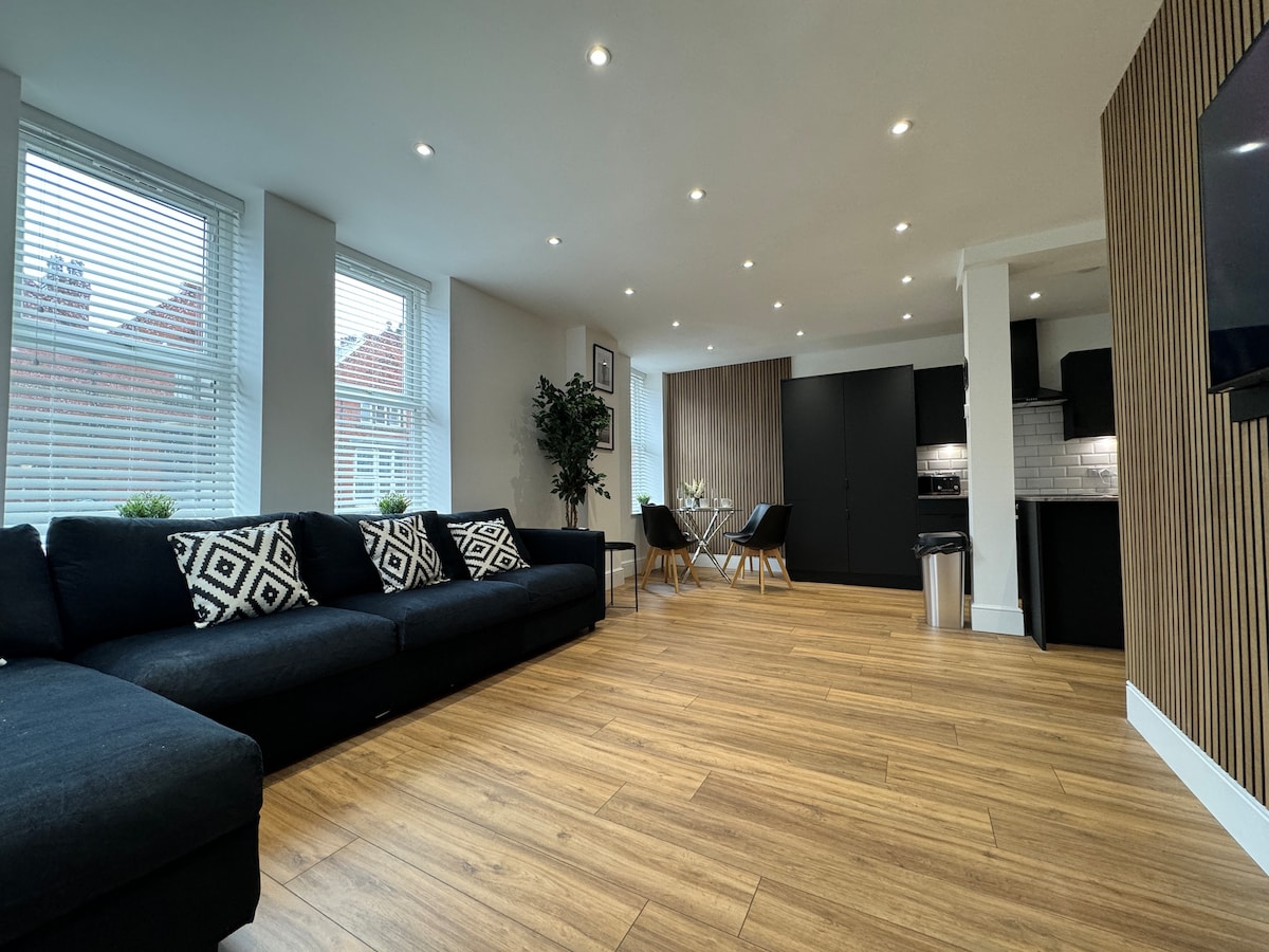 A modern living area features a spacious black sofa adorned with geometric cushions. Natural light flows in through large windows, illuminating the wooden flooring. A dining table with four chairs is visible in the background, adjacent to a sleek kitchen highlighted by dark cabinetry.
