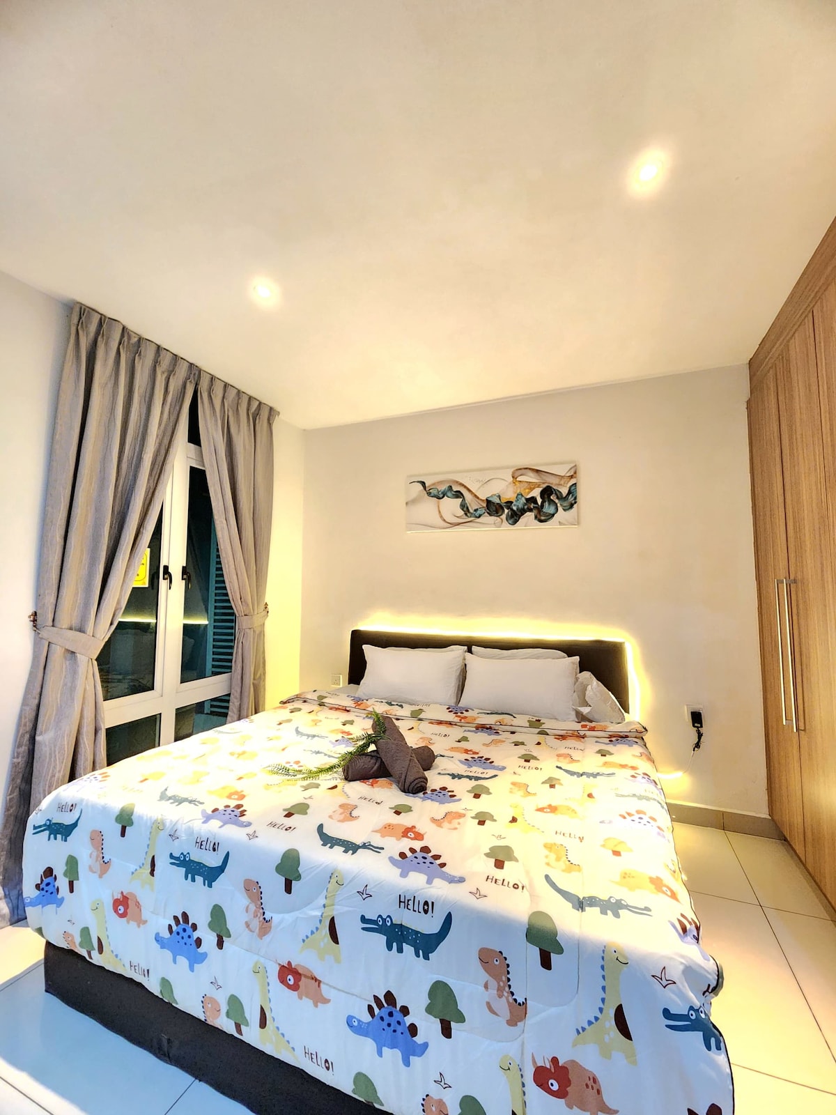 A cozy sleeping area is featured with a king bed, adorned in a colorful dinosaur-themed blanket. Soft light highlights the room's design, complemented by a piece of wall art above the bed. Natural light enters through a window dressed with gray curtains.