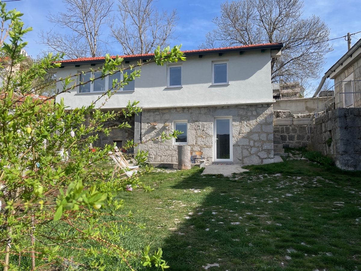 A rustic stone house is surrounded by a grassy yard featuring patches of greenery. Light filters through the branches of nearby trees, and large windows reflect the sky. The back door opens to an inviting outdoor space, creating a serene atmosphere.