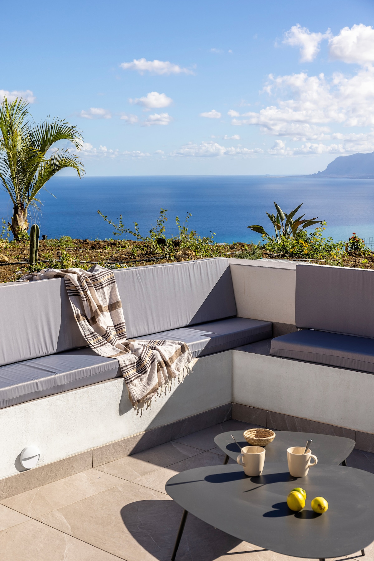 A spacious outdoor seating area features a grey cushion corner sofa with a woven throw, complemented by a small round table. Two cups, a small basket, and fresh lemons are placed on the table. Lush greenery and sweeping ocean views create a serene backdrop.