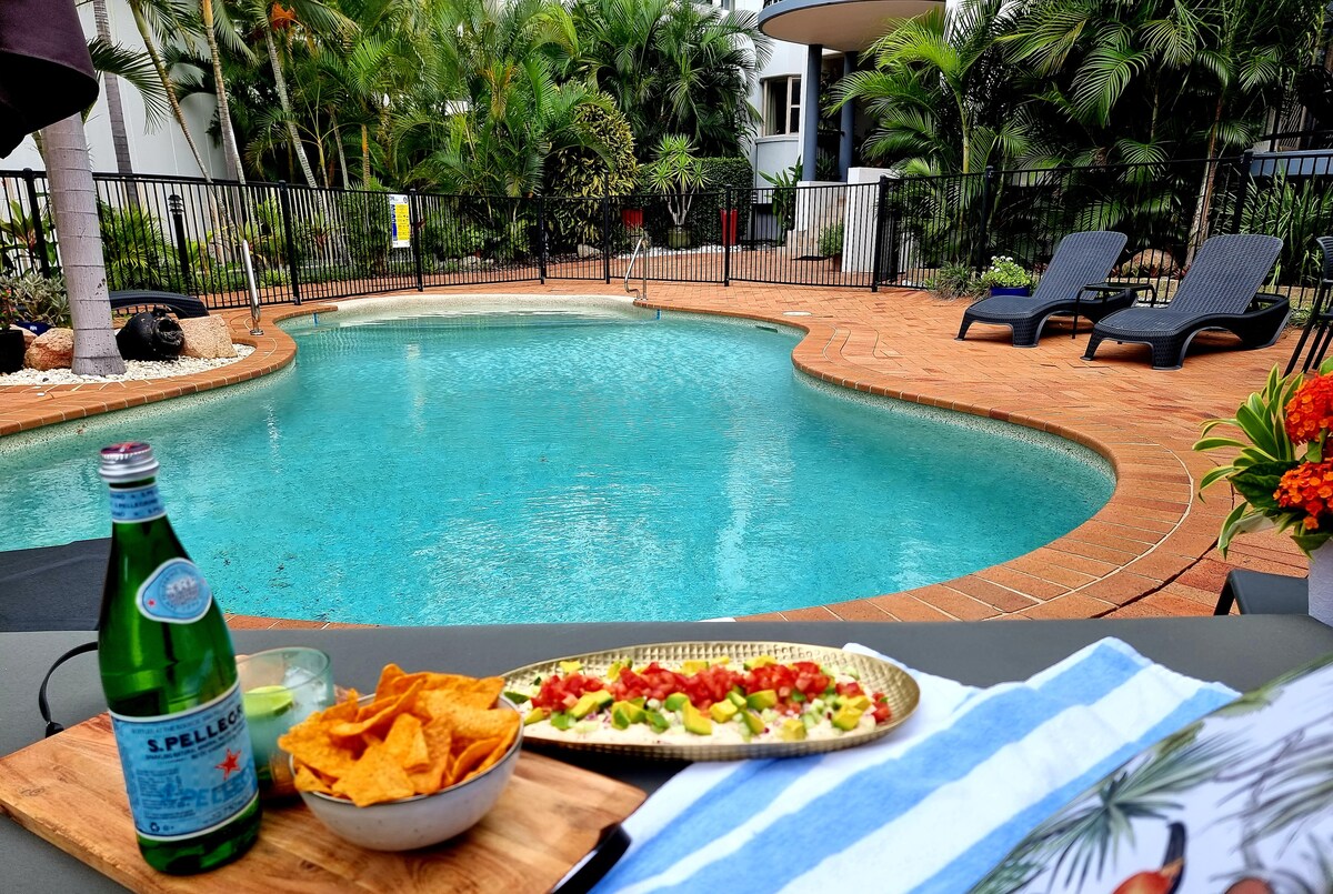 A glistening pool is surrounded by vibrant greenery and loungers, providing a relaxing atmosphere. A wooden platter, featuring snacks and drinks, sits in the foreground, adding to the inviting setting. The pool area is enclosed by a subtle black fence.