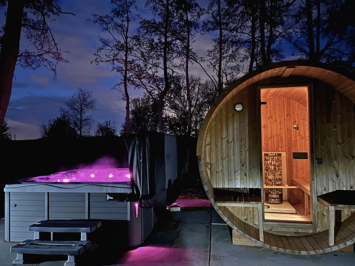 A hot tub with illuminated features is placed beside a wooden sauna. Soft purple lighting enhances the serene atmosphere, while tall trees are visible in the background against a twilight sky.