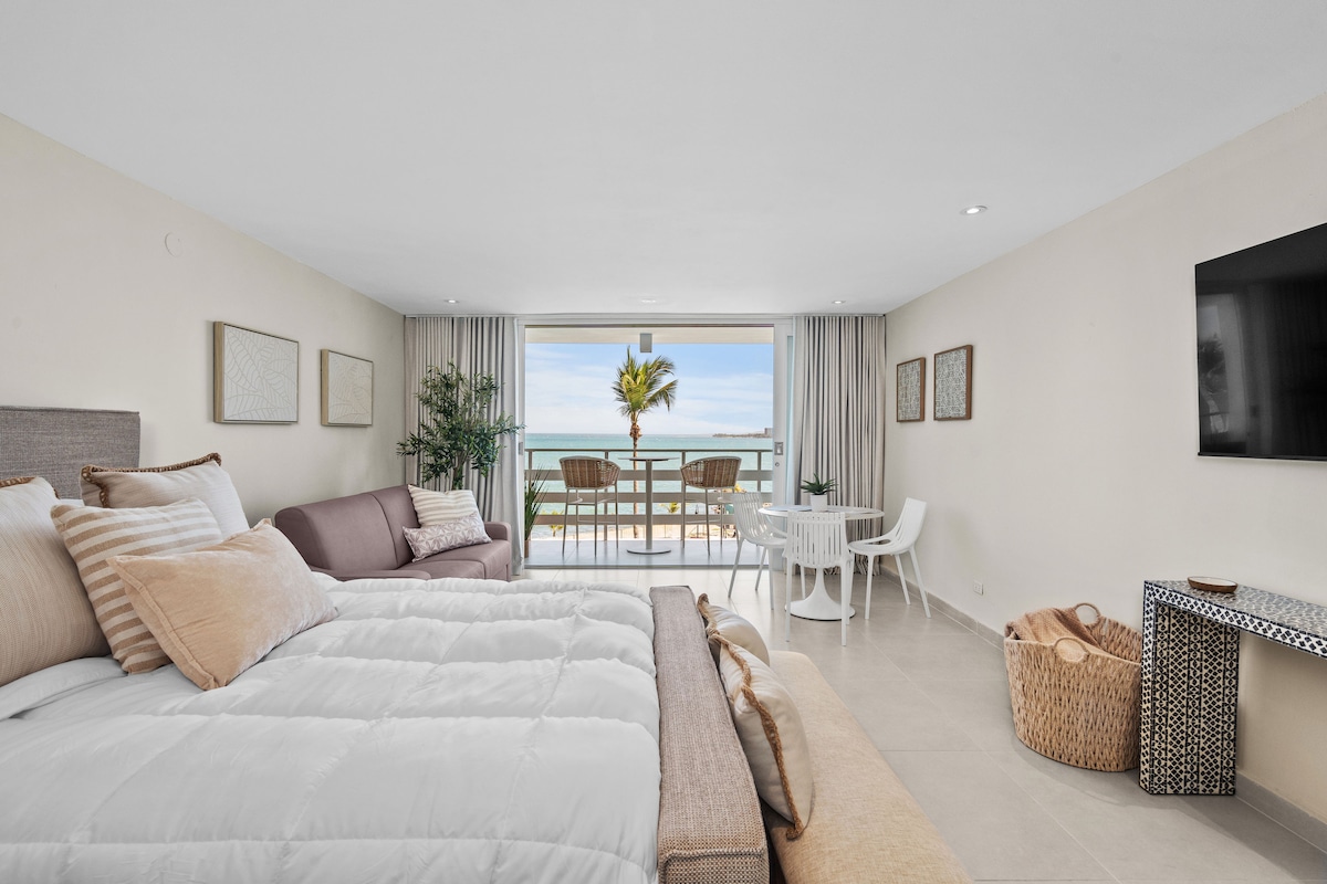 A spacious studio is displayed with a queen-size bed and a sofa bed, both adorned with neutral-toned bedding. A balcony access offers ocean views through glass doors. A small dining table with white chairs and decorative elements enhances the room's inviting ambiance.