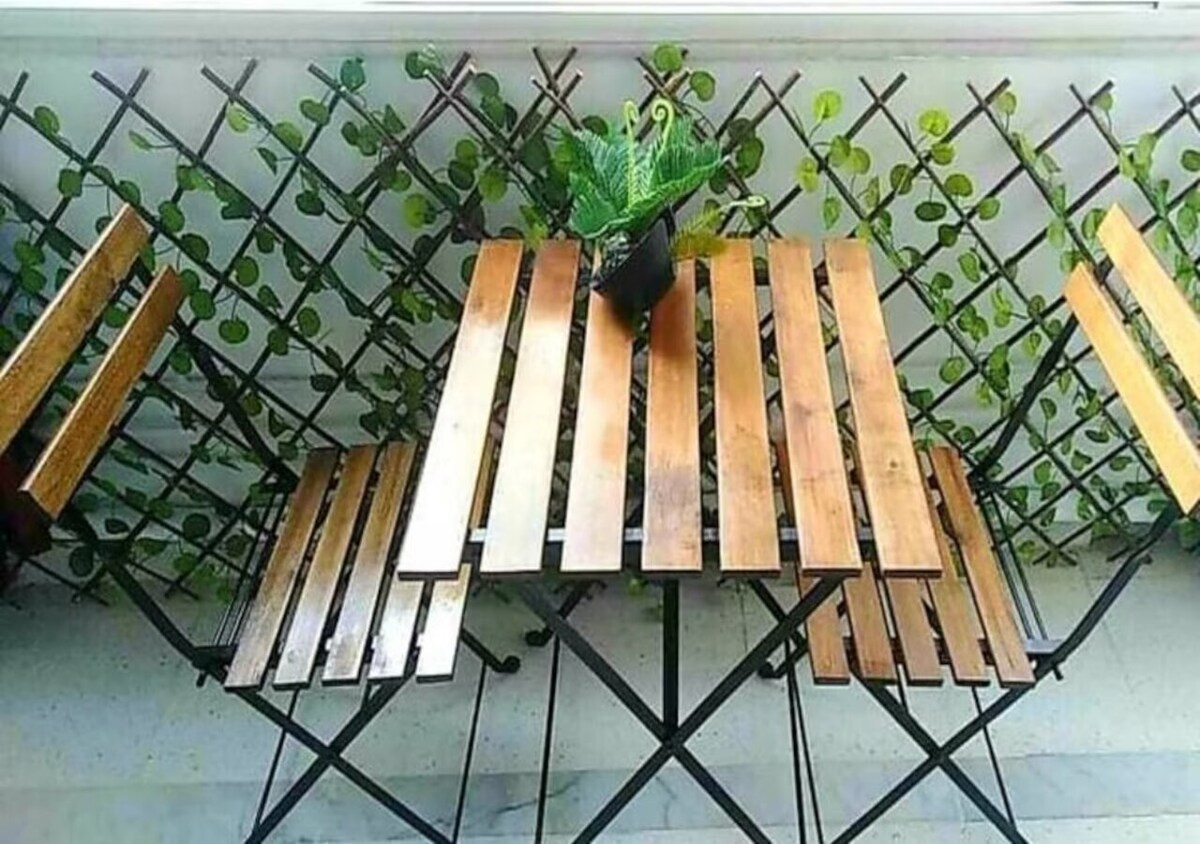 An outdoor balcony area is furnished with a wooden table and two matching chairs. The table features a slatted design, and a small potted plant is placed at its center. Green foliage climbs a trellis in the background, providing a natural touch.