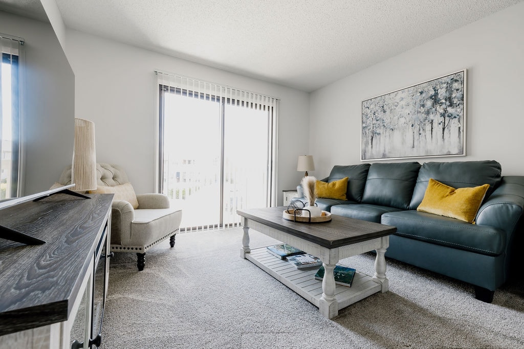 A comfortable living room is showcased with a teal sofa accented by yellow pillows. A wooden coffee table is centered on a plush carpet. Natural light floods the space through glass sliding doors, which lead to an outdoor area.