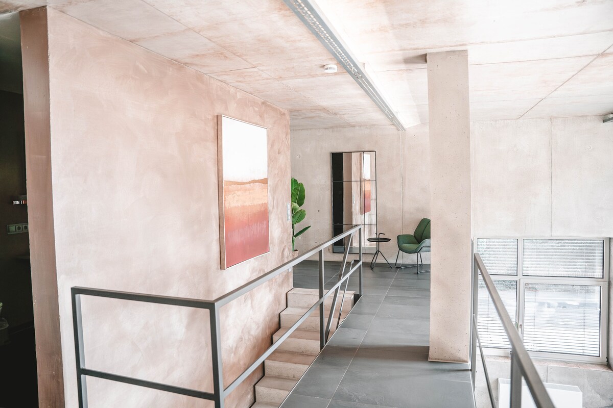 An open gallery area presents a spacious layout with light-colored walls and a polished concrete ceiling. A piece of abstract art adds color to the space, while a minimalist mirror reflects the surroundings. Modern seating and greenery create a relaxed atmosphere.