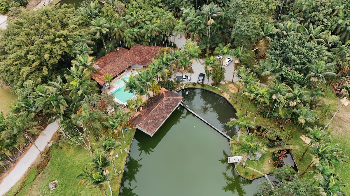 An aerial view captures a spacious property surrounded by lush greenery, featuring a serene lake with a dock. A charming building with a tiled roof is seen near a glimmering pool. Several vehicles are parked, providing a sense of accessibility and convenience.
