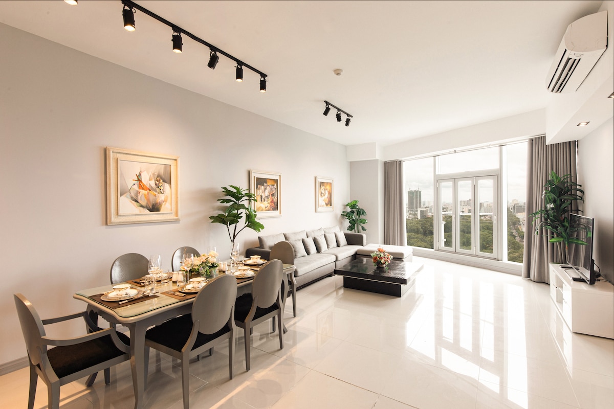 An airy living space features polished floors and large windows, allowing ample natural light. A dining table is set for meals, accompanied by elegant chairs. A stylish sofa anchors the seating area, with tasteful art adorning the walls and greenery subtly included for a fresh touch.