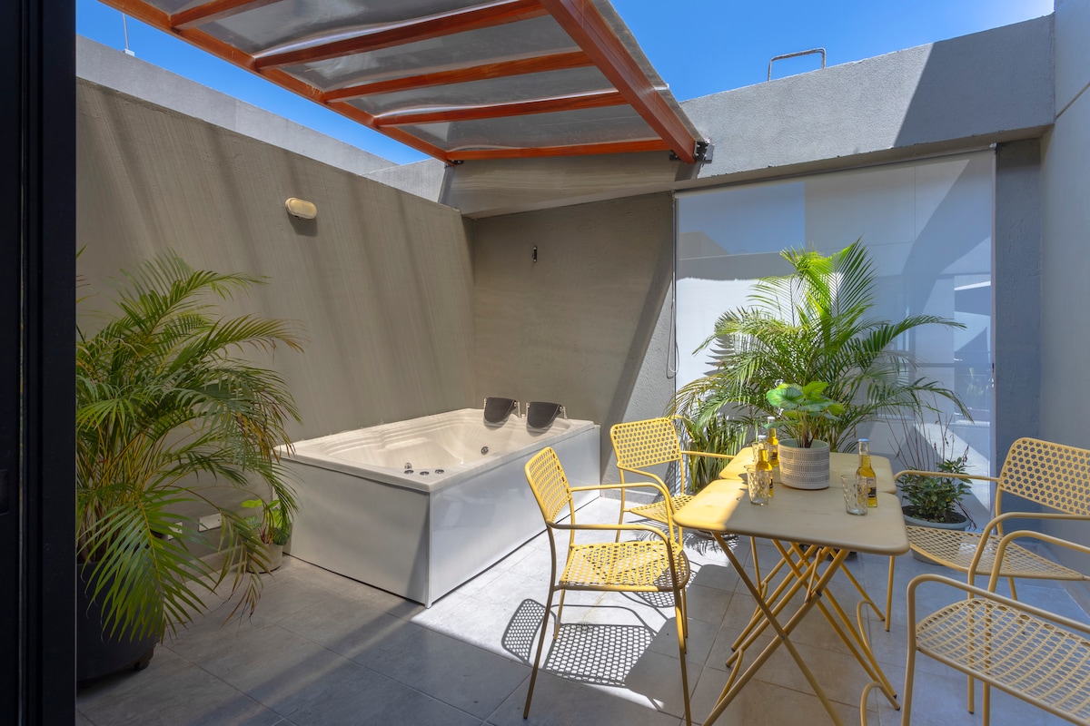 A private terrace features a Jacuzzi alongside tropical plants, providing a relaxing outdoor space. A small dining table with four chairs in a cheerful yellow color is positioned nearby, inviting guests to enjoy meals in the fresh air under a partial canopy.