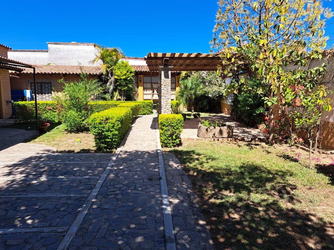 A landscaped outdoor area features stone pathways leading to a welcoming entrance. Lush greenery surrounds the space, including neatly trimmed hedges and a variety of plants. A pergola provides shade, while a few trees and vibrant flowers enhance the tranquil atmosphere.