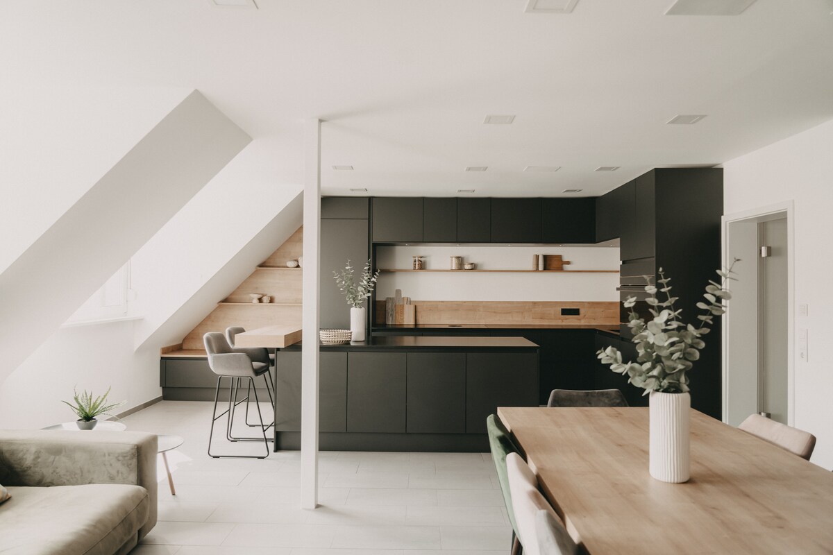 The modern kitchen features sleek black cabinetry and wood accents, creating an elegant contrast. A spacious dining table is complemented by comfortable seating, while a built-in nook offers a cozy area for relaxation. Natural light enters through windows, illuminating the contemporary design.