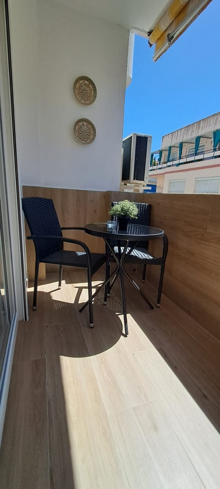 A small balcony is featured, furnished with two black wicker chairs and a round glass table. A potted plant sits at the center of the table, and wooden paneling creates a warm ambiance. Bright sunlight illuminates the space, enhancing its inviting simplicity.