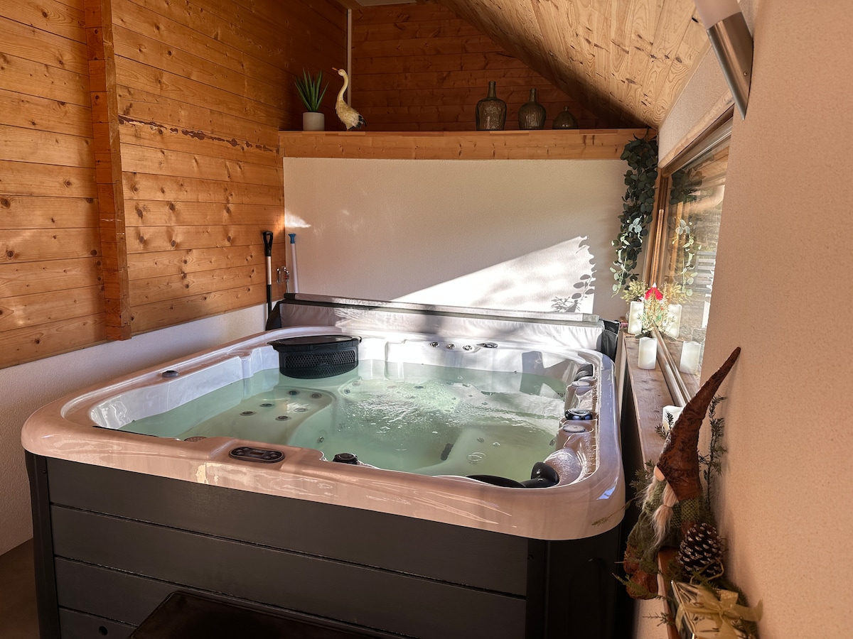 A spacious indoor Jacuzzi is positioned against a wooden wall, reflecting natural light from nearby windows. The area features tasteful decor, including plants and decorative items, creating a serene environment for relaxation.