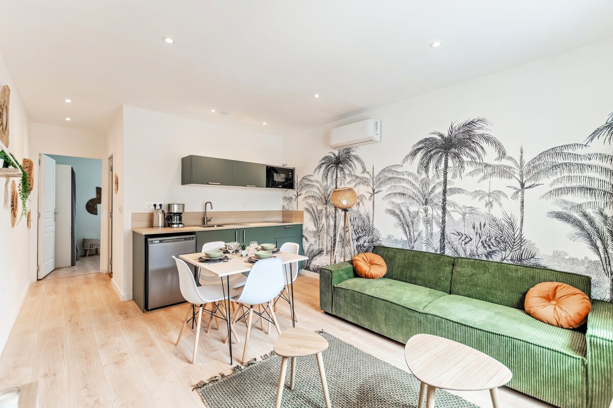 A bright living space is designed with a green sofa and a stylish dining table set for four. A modern kitchen with minimalist cabinetry features essential appliances, while a mural of palm trees enhances the room's tropical feel. Light-colored wood flooring adds warmth.