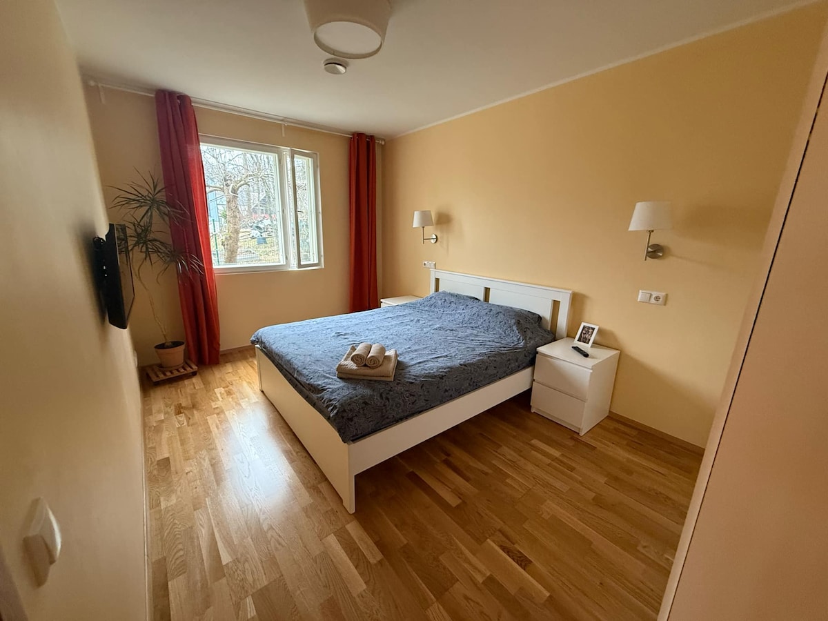 A well-lit sleeping area features a spacious bed with a patterned cover. Two bedside lamps offer soft illumination. A window allows natural light to stream in, while a small plant adds a touch of greenery to the room's warm yellow walls.