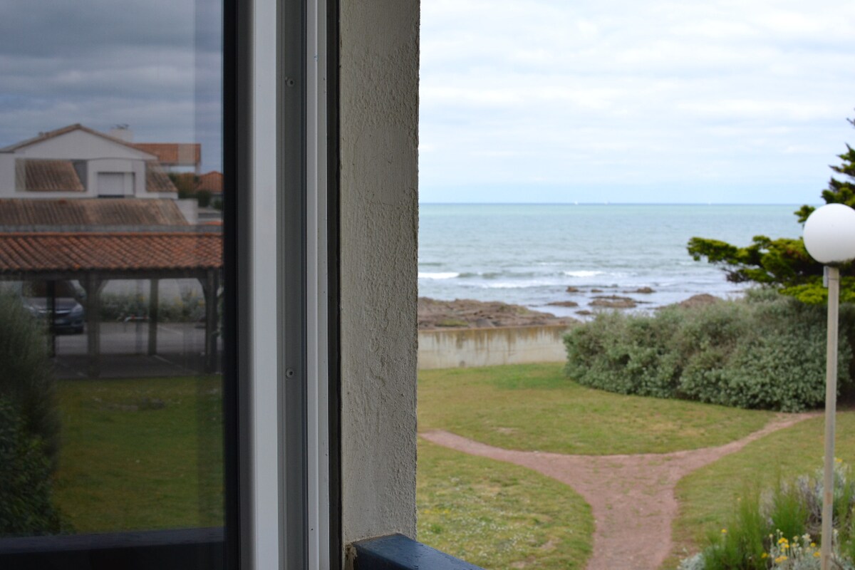 A clear view of the sea can be seen through the window, showcasing gentle waves and a wide expanse of water. The foreground features a well-maintained green lawn bordered by pathways leading to the beach, with shrubs and a light fixture present.