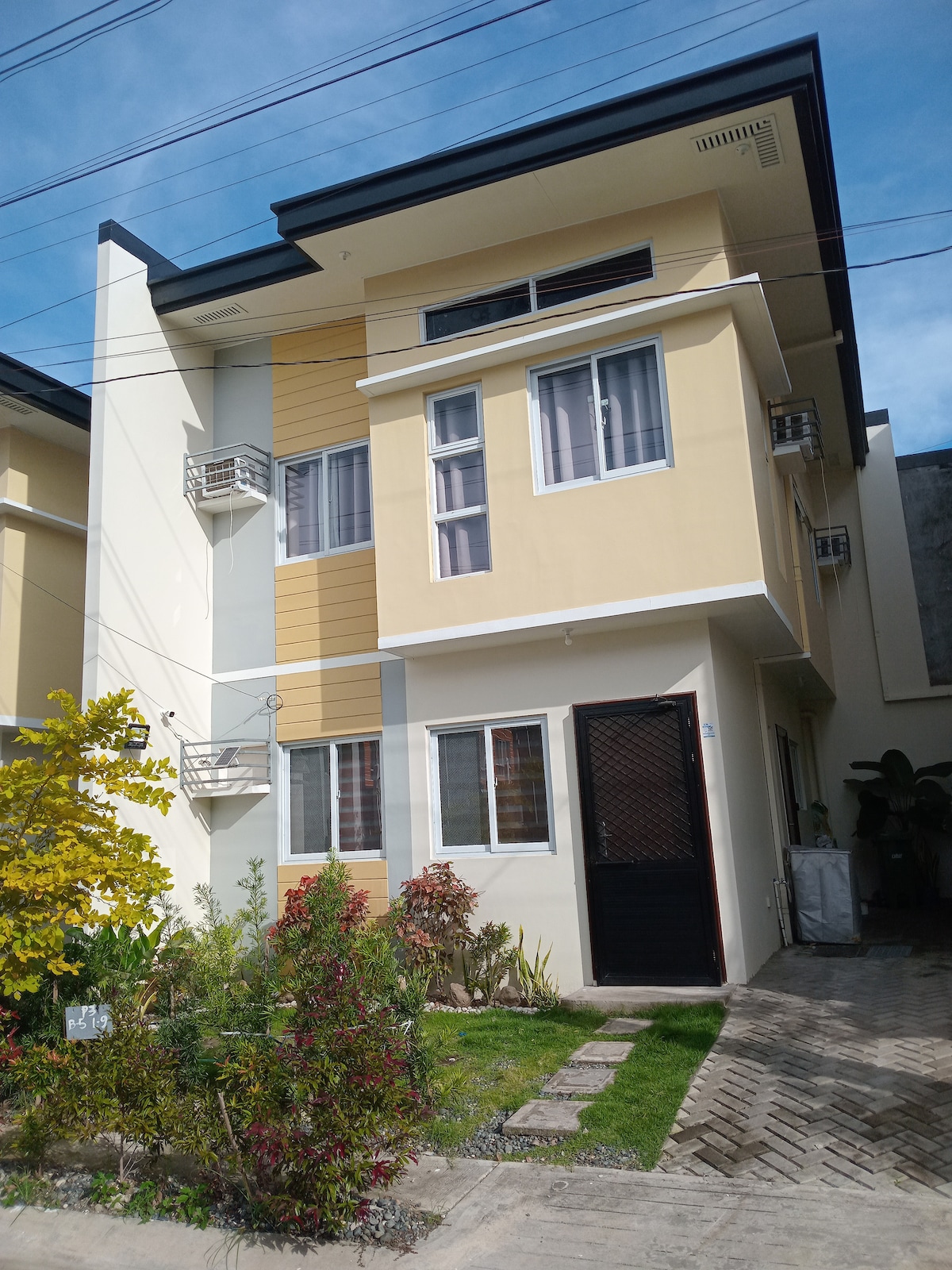 The exterior of a modern, two-story house is displayed, featuring a blend of yellow and white walls. Windows with metal grilles allow natural light in, while a small garden with green plants adds a touch of nature to the entryway. A black door provides access to the property.