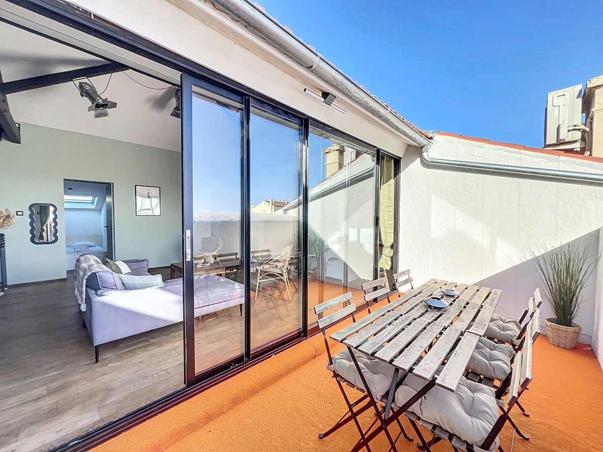 An outdoor terrace is featured with a wooden dining table surrounded by several chairs. Large glass doors open to a spacious interior, allowing natural light to fill the area. The scenic view of the sky above enhances the open atmosphere of the space.