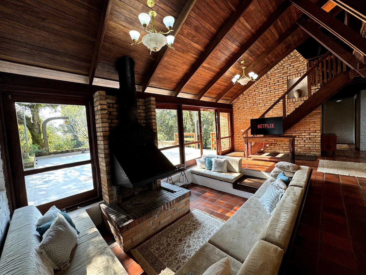 A spacious living area features a large fireplace, wooden beams, and multiple windows with views of the outside. Comfortable seating is arranged around the fireplace, enhancing the inviting atmosphere. A television mounted on the wall offers entertainment options.