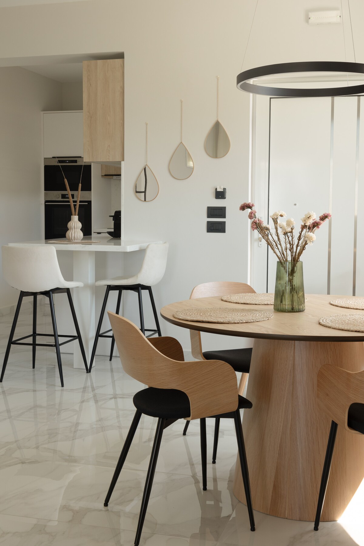 A modern dining area is showcased, featuring a round wooden table with four chairs. A stylish vase with dried flowers sits at the table's center. In the background, a kitchen area and minimalist decor, including mirrors and a pendant light, contribute to the contemporary ambiance.