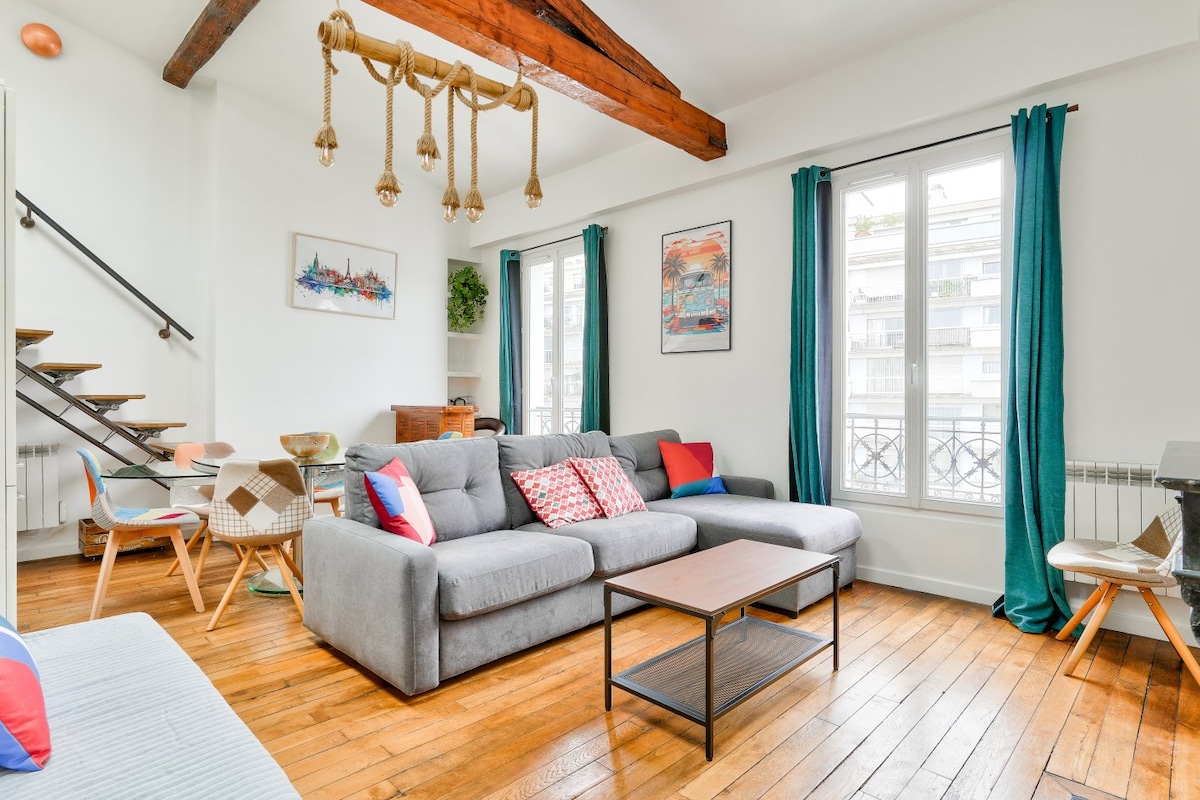 A spacious living area is adorned with wooden beams and features a large gray sectional sofa, accented with colorful cushions. A coffee table sits in front, and large windows are draped with teal curtains, allowing natural light to fill the space.