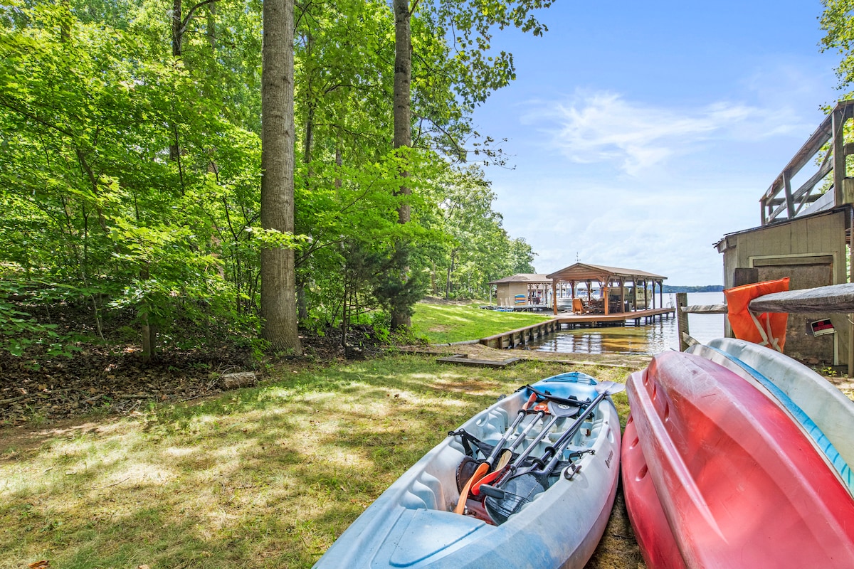 Several kayaks are positioned on the grassy area near the water's edge, surrounded by lush trees. The serene lake can be seen in the background, with a covered sitting area and boat slip nearby, inviting outdoor exploration and relaxation.