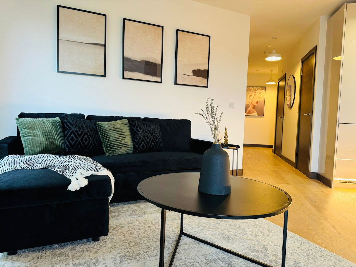 A modern living area is captured, featuring a dark sofa adorned with green cushions and a textured blanket. A round black coffee table is positioned centrally, with a decorative black vase holding dried stems. Minimalist artwork hangs above the sofa, complemented by warm wooden flooring.