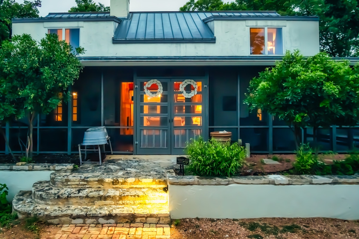 A charming cottage exterior is showcased, featuring a combination of greenery and rustic stone steps. The front porch is framed by large windows and two wreaths adorn the entryway, welcoming guests. Subtle lighting highlights the stone pathway, guiding the way into the home.