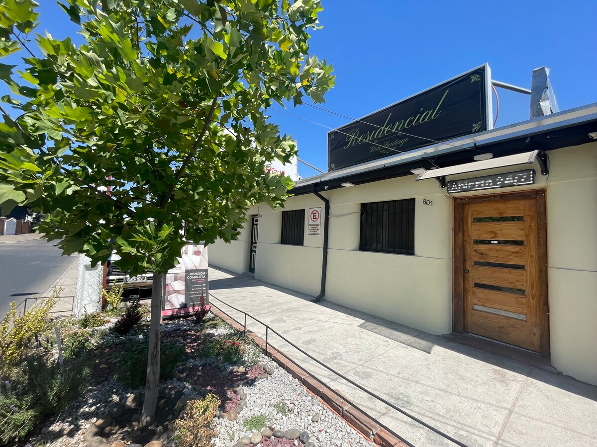 An inviting exterior of the building is highlighted by a well-maintained pathway and modest landscaping. A wooden door is prominently featured at the entrance, and a sign indicates the name of the property. Natural light enhances the overall view of the solid structure.