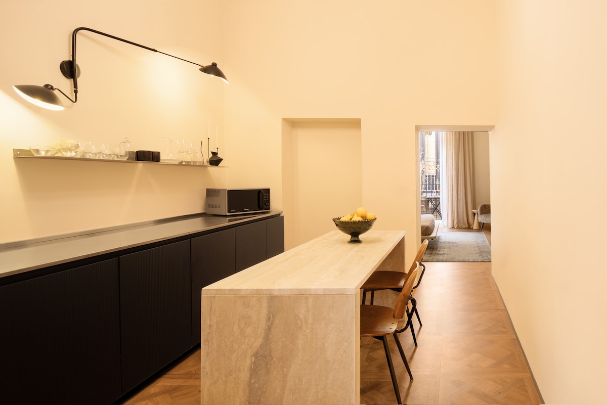 A modern kitchen area is highlighted by a long stone countertop, paired with wooden chairs. Elegant wall-mounted lights illuminate the space, while a doorway in the background leads to a bright living area with natural light filtering through sheer curtains.