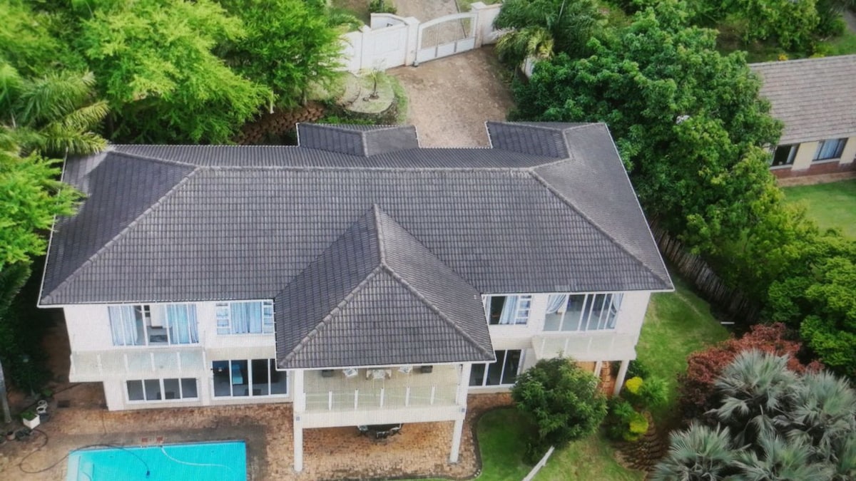 An aerial view of a spacious two-story house is presented, surrounded by lush greenery. The roof features multiple peaks, contributing to the architectural design. A clear swimming pool is visible in the backyard, complementing the natural landscape of trees and well-maintained lawn.