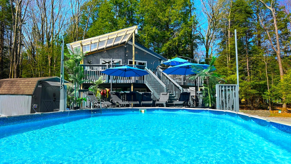 A heated pool is surrounded by lush greenery and a spacious deck. The home features a modern design with large windows and a covered balcony. Umbrellas provide shade for lounge chairs situated around the pool, creating a relaxing outdoor space.