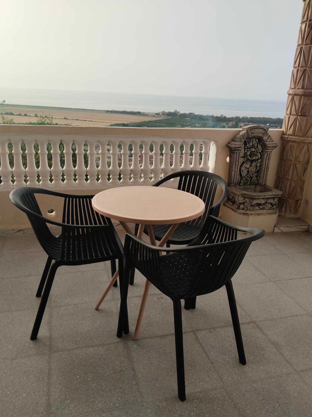 A small round table with three black chairs is situated on a terrace. The sea view is visible in the distance, complemented by a surrounding decorative railing. The flooring is tiled, contributing to the overall outdoor setting.