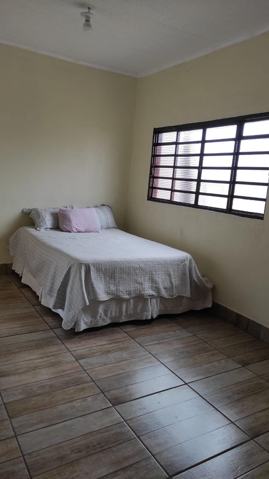 A simple bedroom is presented with a single bed dressed in a light-colored coverlet. A pink accent pillow is placed on the bed. Natural light filters through a grid-patterned window, and the tiled floor extends throughout the room.