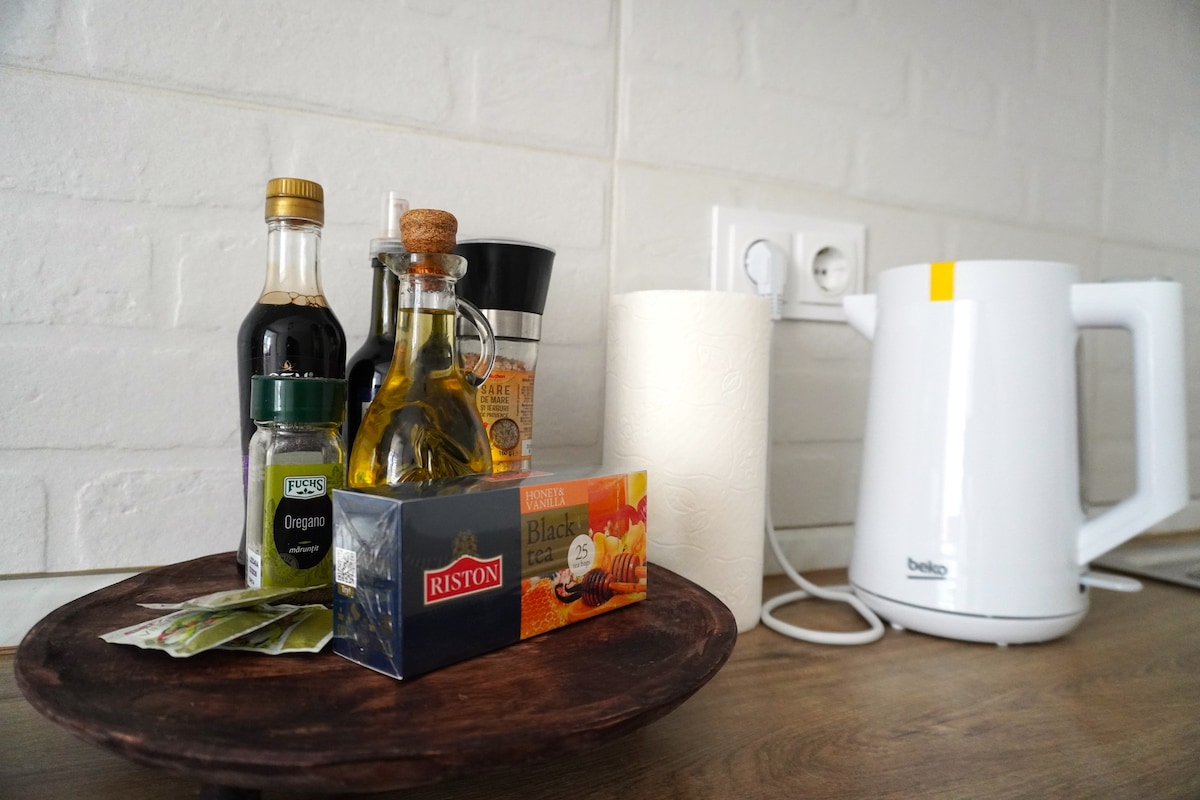 A wooden platter displays various condiments, including olive oil, vinegar, and spices, alongside tea and a roll of paper towels. A kettle is positioned nearby, with a power outlet visible in the background.