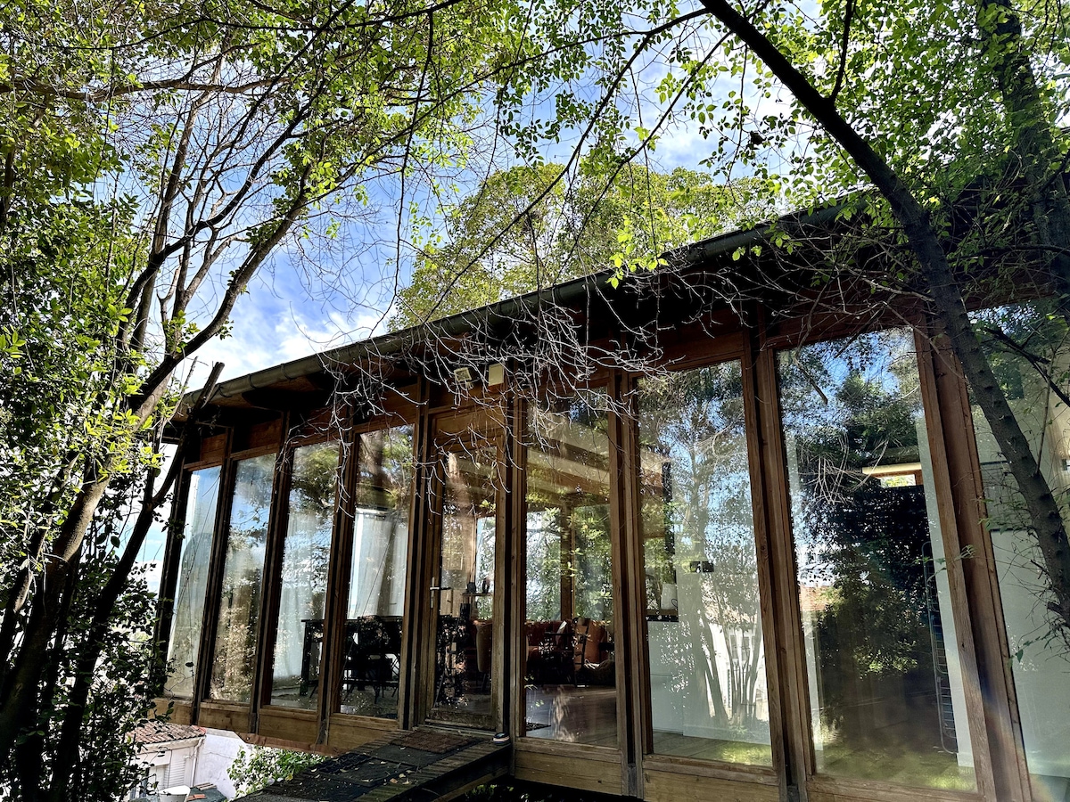 The exterior of the house is encased in large glass panels, surrounded by lush green trees. Natural light filters through, highlighting the wooden structure and offering a seamless transition between indoor and outdoor environments.
