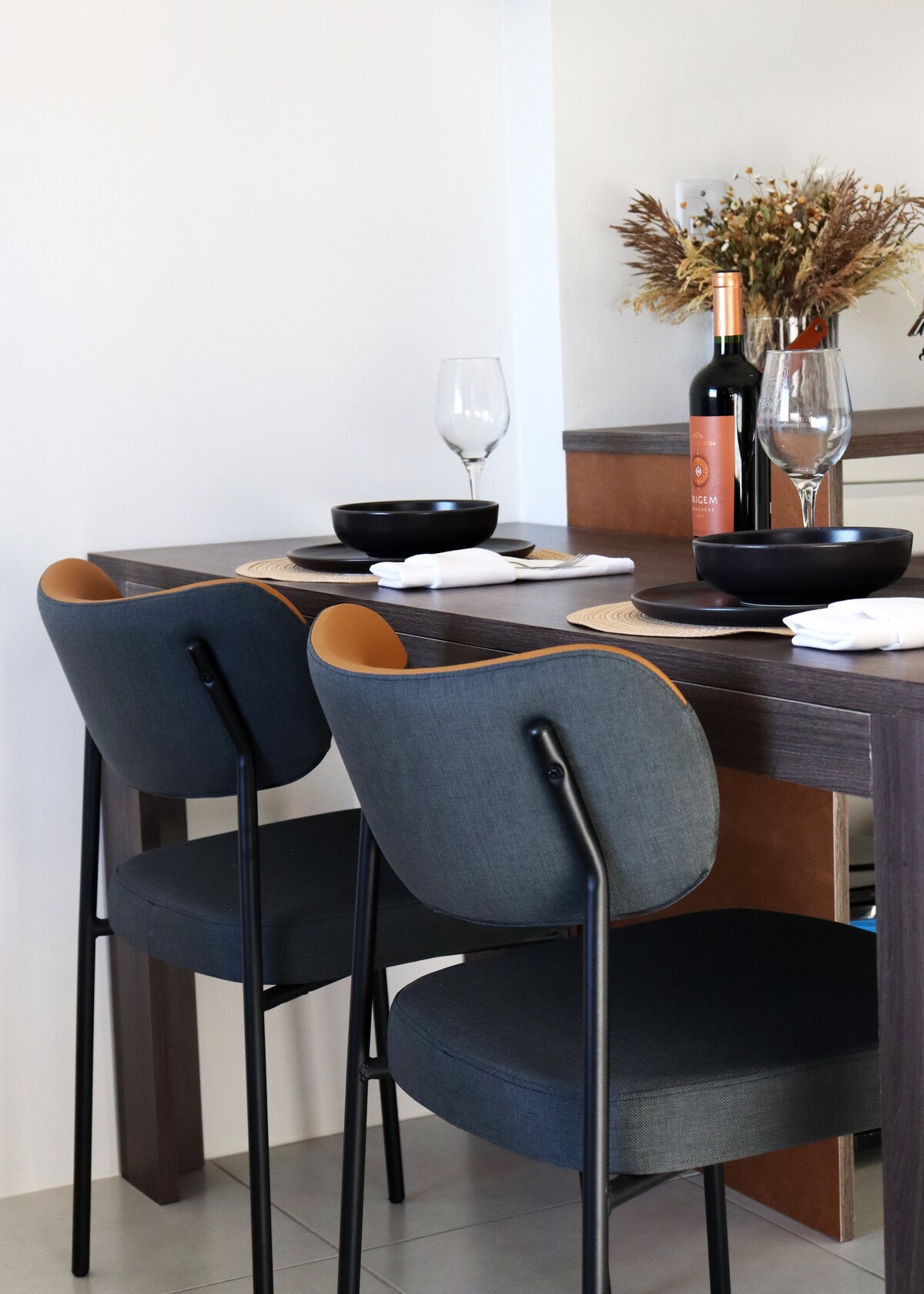 A dining area is featured with two modern chairs, showcasing a dark wood table set for a meal. Plates and glasses are arranged neatly, with a bottle of wine placed subtly in the background, bringing a touch of elegance to the space.