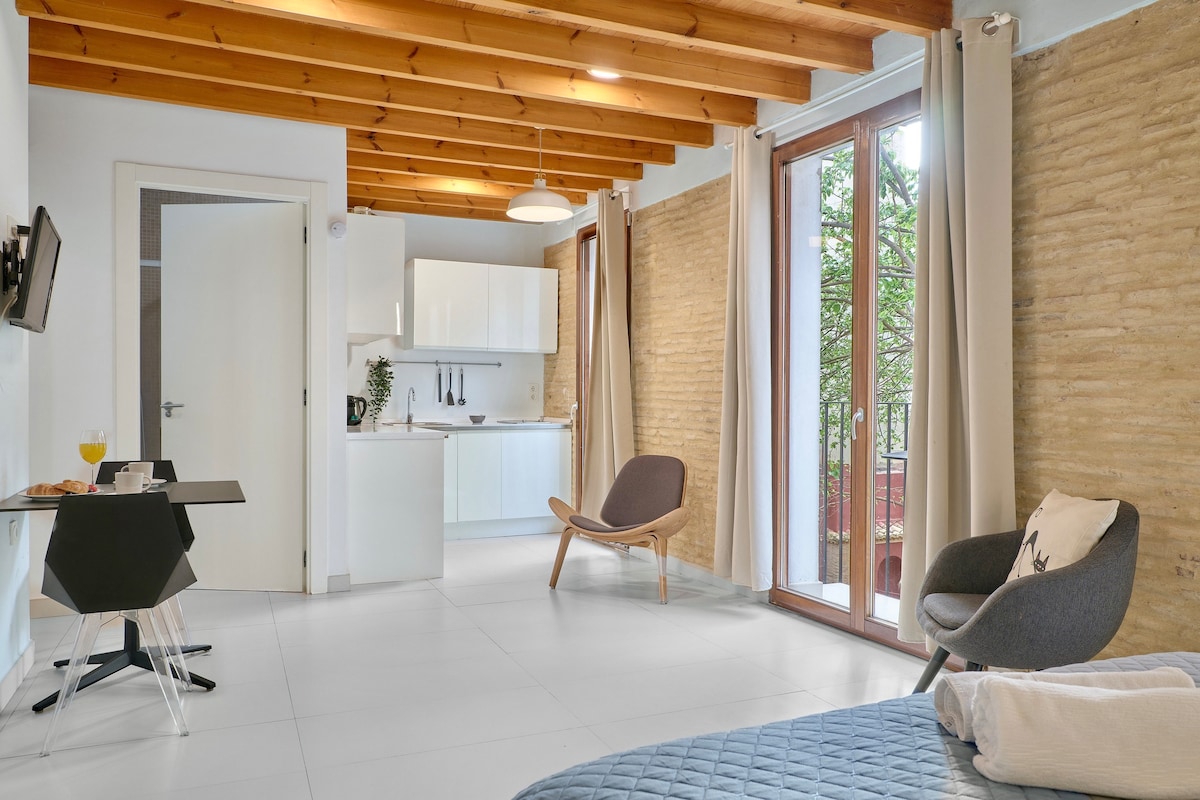 An airy loft space features a corner kitchenette equipped with essential appliances. Natural light filters in through large windows, illuminating the minimalist decor. A cozy seating area includes a modern chair and a small table, while exposed wooden beams enhance the room's charm.