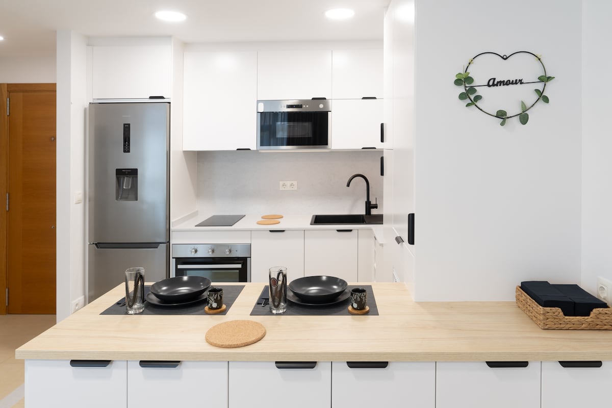A modern kitchen is presented with sleek white cabinetry and a light wooden countertop. Equipped appliances include a refrigerator, oven, and a microwave. Black dining ware is set on the countertop, alongside a natural fiber placemat. A decorative heart-shaped wall piece adds a personal touch.